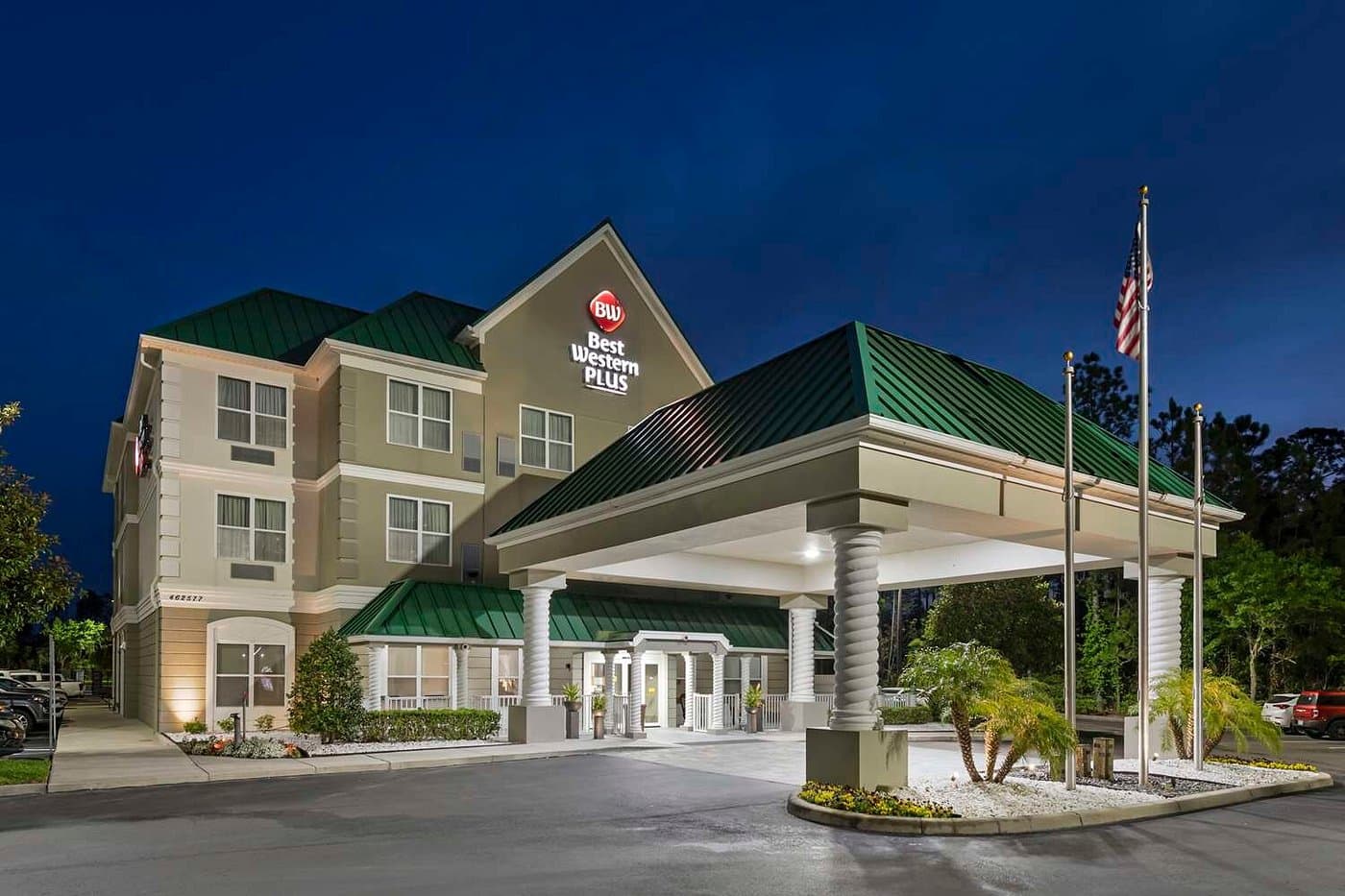 The Best Western Plus First Coast Inn & Suites at dusk, a three-story building with a green roof, illuminated sign, and a columned covered entrance with flags out front