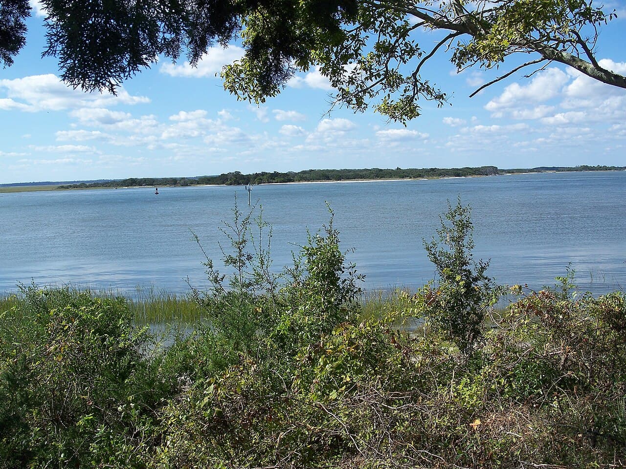 A calm stretch of the Amelia River at Fernandina Beach, a wooded far shoreline and channel markers on the water, framed by green foliage under a blue sky with scattered clouds