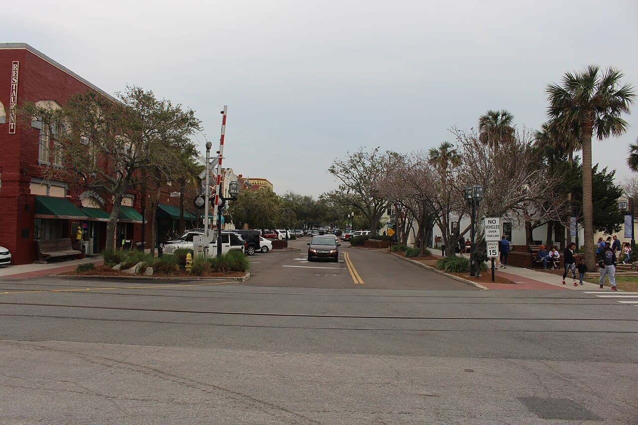 Centre Street in downtown Fernandina Beach looking east, brick storefronts with a RESTAURANT sign, palm and street trees, a railroad crossing in the foreground and people on the sidewalk